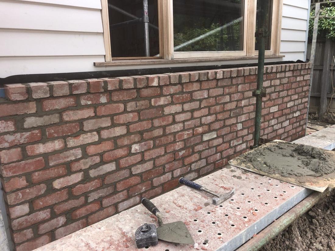 Freshly laid reclaimed brick base wall below a timber window, with trowel and hammer on the scaffold